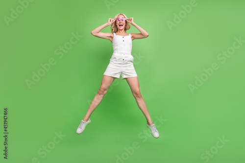 Full size photo of young excited smiling girl jumping summer vacation wear white overall isolated on green color background © deagreez