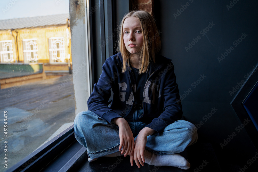 portrait of a girl with blue eyes and blond hair sitting near the window. Looking at camera. Alone. Sad face portrait of a girl with blue eyes and blond hair sitting near the window. Looking at camera. Alone. Sad face