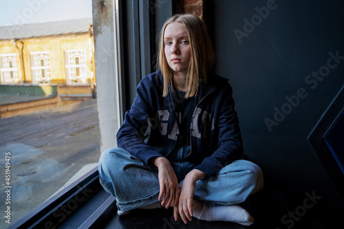 portrait of a girl with blue eyes and blond hair sitting near the window. Looking at camera. Alone. Sad face