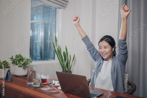 A medium shot of an Excited young adult Asian woman, doing video call conference meetings over a laptop computer on a wooden table. She is enjoying her success.