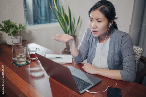 A medium shot of an angry young adult Asian woman, in a grey cardigan and white t-shirt inside doing video call over a laptop computer on a wood table. She strongly disagrees with the conference.