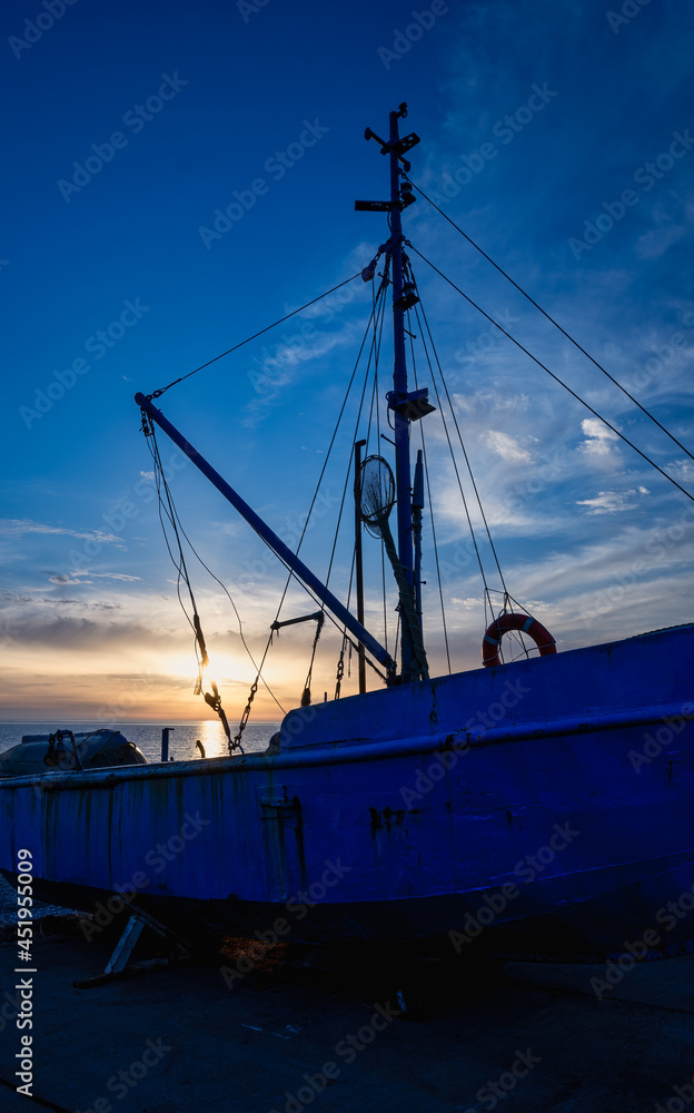 Blue rusting fishing boat on the seashore at sunset. Yellow sun rays are reflected on the water surface. The concept of fishing, transportation, travel and seascapes.