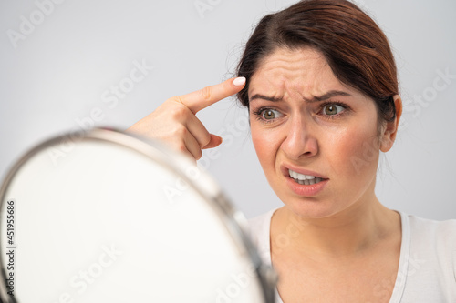 Bild auf Leinwand Caucasian woman looks at herself in the mirror and is upset because of facial wrinkles on her face