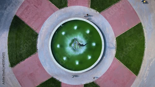 Monument to astronaut and a fountain in middle of a symmetrical circular square. Neatly mown lawn is separated by paving paths. Favorite place for walks of townspeople. Drone moves up quickly.