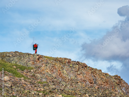 Photographer with a large backpack takes pictures of a beautiful mountain landscape on the edge of a cliff under blue sky. Dangerous mountains and abyss.