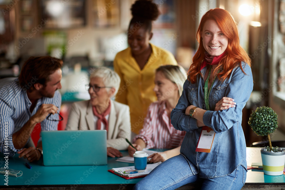 young adult redhead female posing at work in front of group of working people, smiling, looking at camera.