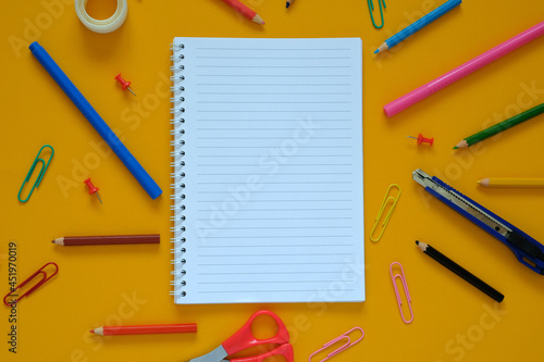 Flat lay. Yellow background. In the center lies a notebook on a spiral, around the stationery: paper clips, pencils, scissors. September 1, knowledge day, business.