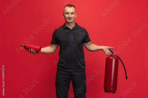 Fire safety. Portrait of young smiling man with extinguishers posing over red background. photo