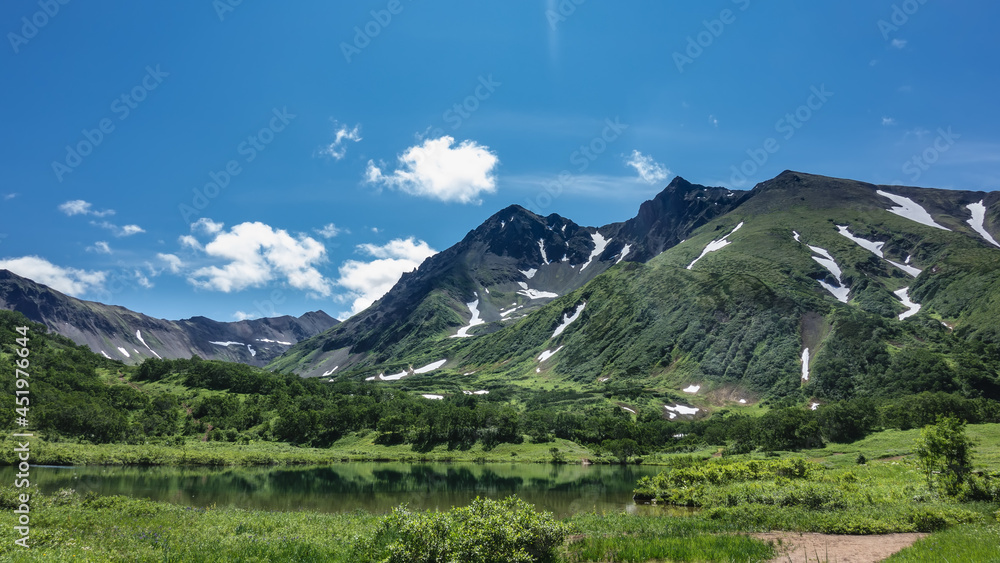 A picturesque lake in the valley, next to a mountain range. There is green vegetation on the banks, snow patches on the slopes. Reflection on the water surface. Blue sky. A summer day. Kamchatka