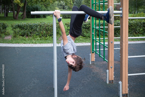 Boy doing exercise upside down on crossbeams on outdoor playground against the backdrop of green bushes photo