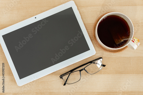 Tablet, eyeglasses and tea cup on wooden office desk.
