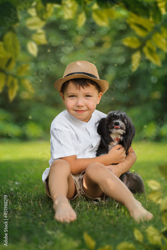 A little white boy in a hat and a white shirt is sitting on a green summer lawn with his little black dog