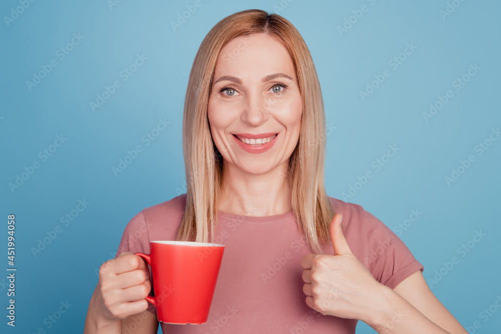 Photo of cheerful woman holding mug of coffee show thumb-up like cool advert isolated over blue color background Photo of cheerful woman holding mug of coffee show thumb-up like cool advert isolated over blue color background