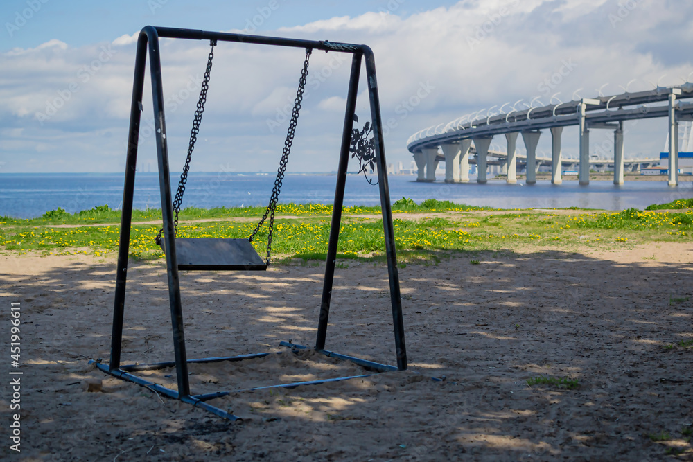 Fototapeta premium Empty swing swayed by the wind in shade of trees on the coastline seashore near of a big rounded Controlled-access motorway highway bridge, the road passing over the sea bay coast