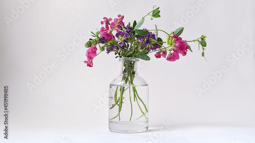 Tablou pe pânză A bouquet of pink and lilac wildflowers in a small transparent vase on a white b