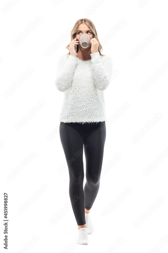 Walking young woman in cozy home wear talking on t he phone looking up holding coffee. Full body portrait isolated on white background Walking young woman in cozy home wear talking on t he phone looking up holding coffee. Full body portrait isolated on white background
