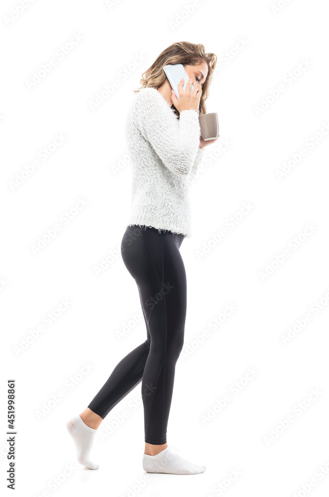 Side view of young woman in home wear white sweater walking talking on the phone with coffee mug. Full body portrait isolated on white background Side view of young woman in home wear white sweater walking talking on the phone with coffee mug. Full body portrait isolated on white background