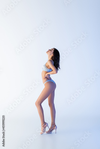 Fototapeta Naklejka Na Ścianę i Meble -  side view of young woman in lace lingerie posing with hand on hip on blue