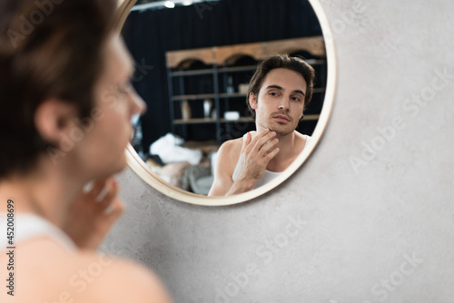 young man touching unshaven face while looking in mirror photo