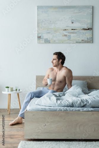 shirtless man looking away while sitting on bed with cup of coffee
