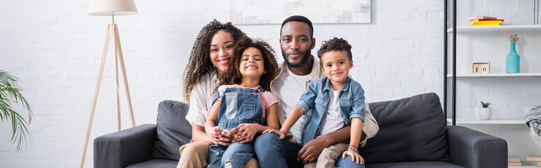 joyful african american family smiling at camera on sofa at home, banner