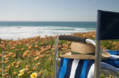 Straw sun hat and towel on empty directors chair sitting on a bank full of flowers overlooking the beach photo