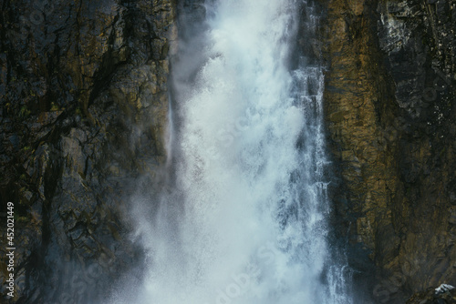Atmospheric minimal landscape with vertical big waterfall on rock mountain wall. Powerful large waterfall in dark gorge. Nature background of high vertical turbulent falling water stream on wet rocks.