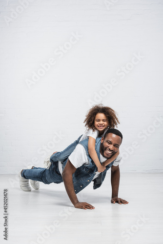 cheerful african american girl piggybacking happy dad doing push ups on grey