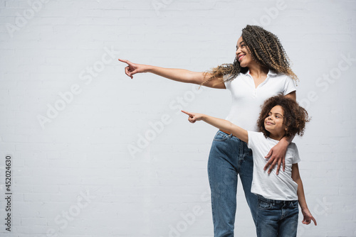 smiling african american mother and daughter looking aside and pointing with fingers on grey
