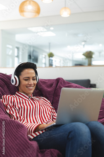 Smiling caucasian female business creative wearing headphones, lying on sofa and using laptop photo