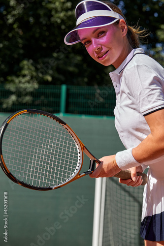 Confident Young Caucasian Lady In White Uniform And Cap Holding Tennis Racket In Hands, Smiling, Motivated Sportswoman On Hardcourt At Summer Day. People Lifestyle, Fitness, Sport Concept © Roman