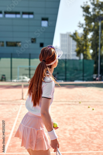 Rear view on pretty young woman in sexy sportwear playing tennis in city park at hot summer day. Redhead female tennis player with racket, enjoying game, looking at side. Sport, healthy lifestyle © Roman