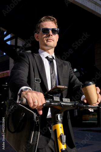 Portrait of business person with electric scooter outside in front of modern business building, looking directly, wearing formal clothing and sunglasses, enjoying free time after working day