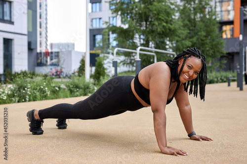 Fitness lifestyle. Young smiling fat overweight african woman in black tracksuit doing push ups, side view sporty young lady at summer day in sport ground. Healthy life concept. Copy space
