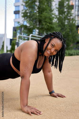 Fitness lifestyle. Optimistic fat overweight african woman in black tracksuit doing push ups, side view sporty young female at summer day in sport ground. Healthy life concept. Copy space. sport