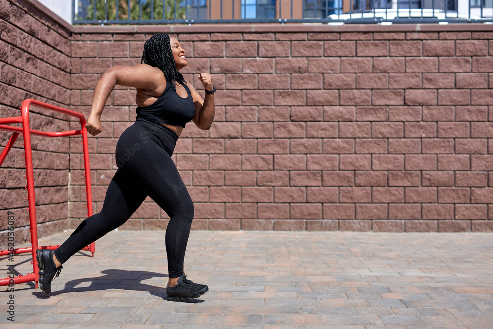 African fat woman jogging during cardio workout for burning calories outdoors in city at day time, side view, copy space. Young obese lady in black sportive tracksuit is running forward, focused. - obrazy, fototapety, plakaty 