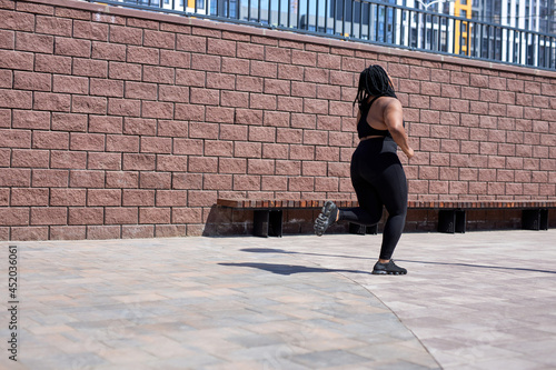 An obese young african female jogging outdoors. Fat beautiful pretty female in black tracksuit is engaged in fitness for weight loss in city. Side view on beautiful chubby woman runs on summer day. © Roman