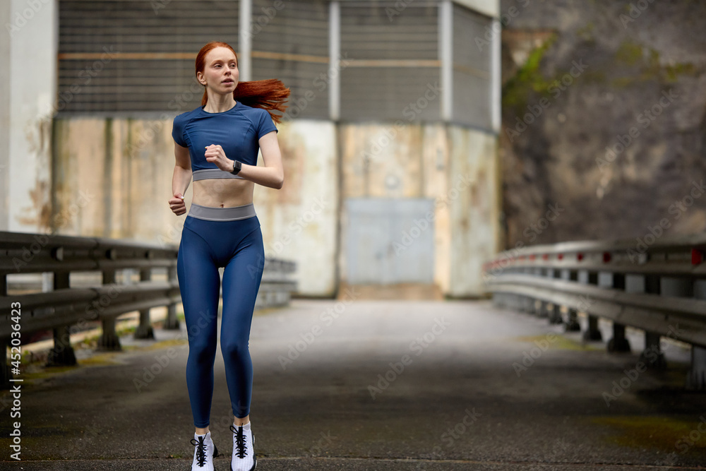 Young slim redhead woman warming up on bridge before workout, running. Beautiful young fit fitness sport model jogging alone, side view. healthy lady in blue sportive clothes. motivation concept