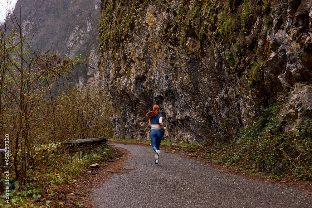 Fototapeta premium Good-looking caucasian woman exercise by running on the road near forest mountains, side view, red-haired lady is dressed in sportive clothes, copy space. healthy lifestyle, sport concept. rear view