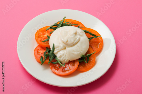 Delicious fresh cheese made from cream and milk - burrata. Italian tender cheese with tomato and greens salad on a bright pink background
