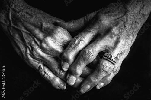 A close up black and white image of an elderly woman's arthritic hands. She is wearing a diamond ring and her knuckles are swollen. Her hands are gently placed on each other. Her nails are painted. 