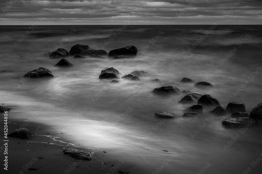 Fototapeta premium A moody black and white image of waves rolling into a rocky shore. A long exposure created a misty, dreamy feeling coupled with dark clouds. The image creates a feeling of peace and tranquility.