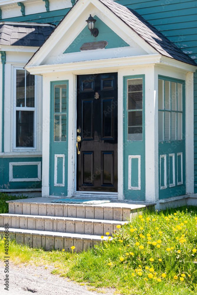 The exterior of a vintage colonial style teal green wooden building with white decorative trim. There's an old black wood door with brass handles and a knocker. Three steps lead up to the building. 