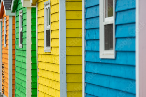 A row of small colorful painted huts or sheds made of wood. The vibrant exterior walls are colorful. Painted yellow  blue  red  and green with small white double hung windows in the centers.