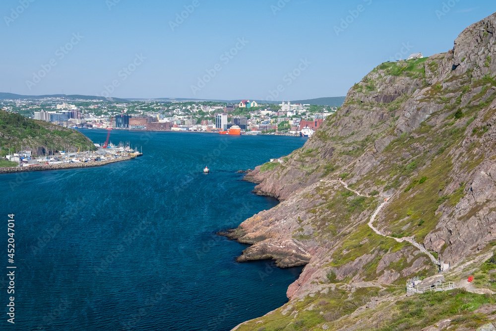 A footpath, hiking trail or path along a hillside. The cliff is rocky with grass patches. The city of St. John's, Newfoundland, is in the background on a sunny day. The sky is bright blue. 