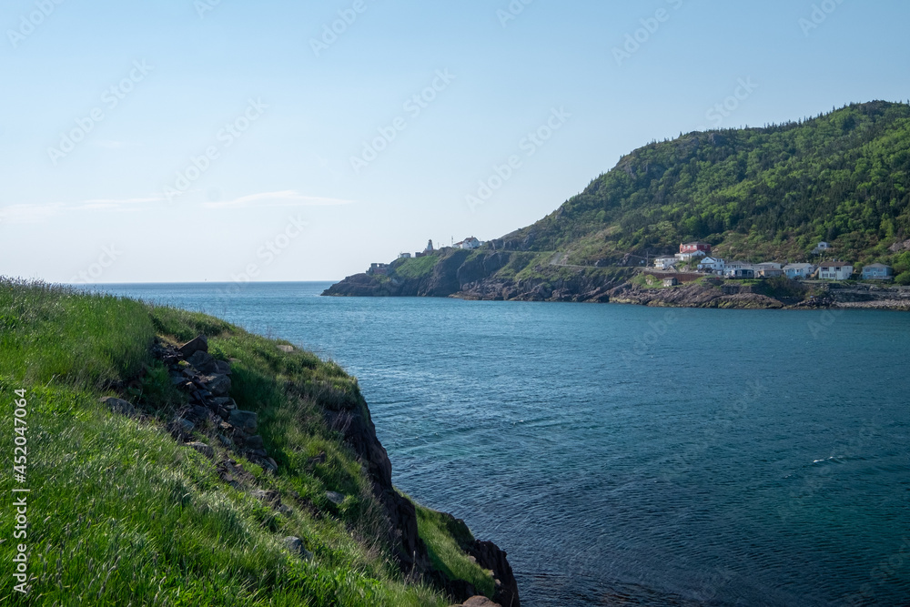 A footpath, hiking trail, or path along a hillside next to the ocean. The cliff is rocky with grass patches. St. John's, Newfoundland, is in the background on a sunny day with small wooden houses.