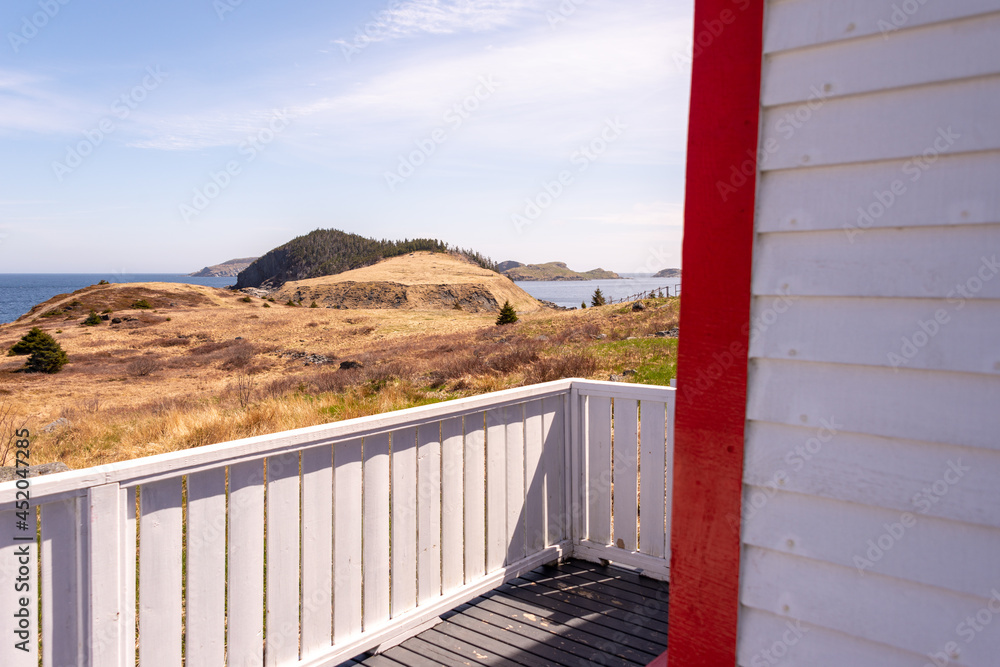 Fototapeta premium The exterior of a vintage white wooden building is covered in narrow white cape cod clapboard with vibrant red wood trim. There's a view of hills, fields, and the blue ocean off the balcony. 