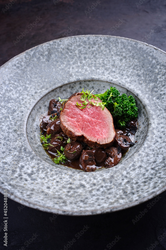 Modern style traditional fried dry aged angus beef filet medaillons natural with mushrooms and baby broccoli in red wine sauce served as close-up on a design plate Modern style traditional fried dry aged angus beef filet medaillons natural with mushrooms and baby broccoli in red wine sauce served as close-up on a design plate