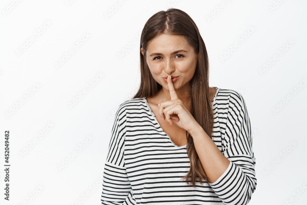 Coy young woman having secret, hushing and smiling devious, be quiet, taboo gesture, making shh sound, standing over white background Coy young woman having secret, hushing and smiling devious, be quiet, taboo gesture, making shh sound, standing over white background