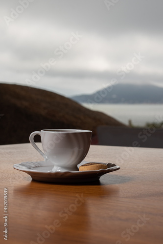 Small white coffee set with a biscuit on the side, on a wooden table. In the background a sea and a mountain landscape on a cloudy day.